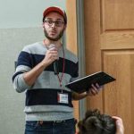 Julian Gonzalez in a hat and striped sweater, speaking into a microphone.