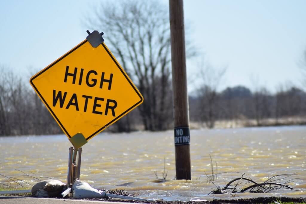 A yellow sign says "high water" in front of a flooded roadway backed by leafless trees.